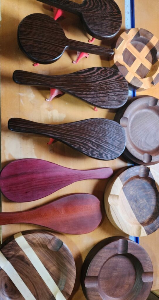 Handcrafted wooden paddles and ashtrays in progress, showcasing various exotic and domestic wood species with intricate finishes and unique designs.