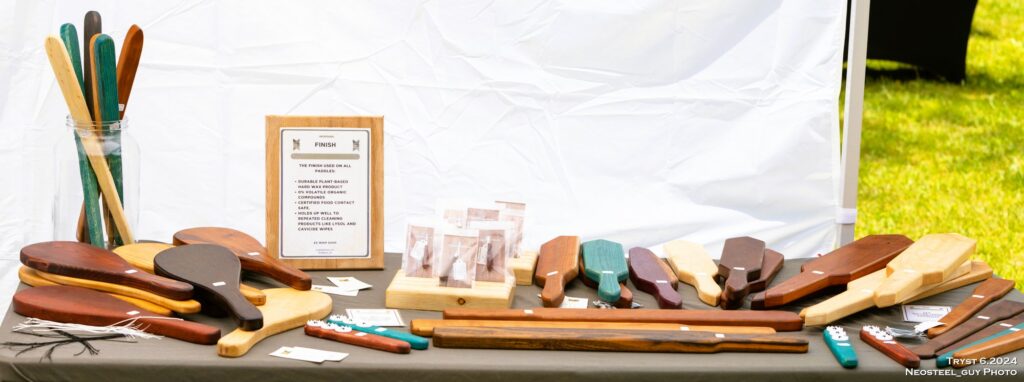 A vendor table at an outdoor event displaying handcrafted wooden paddles, canes, paracord whips, and other finely crafted wood products.